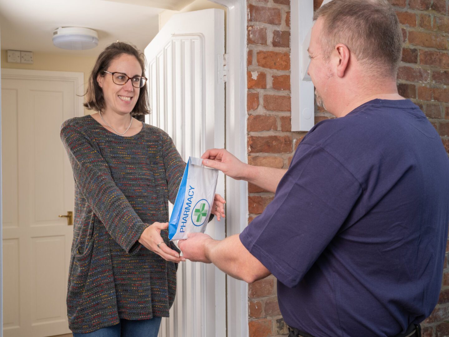 A man hands a pharmacy prescription package to a woman at the doorway of a brick house, capturing a moment of medical prescription delivery in a residential setting.